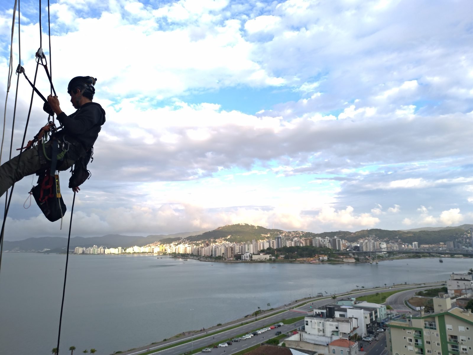 Profissional em acesso por cordas na fachada de edifício, com a baía de Florianópolis ao fundo