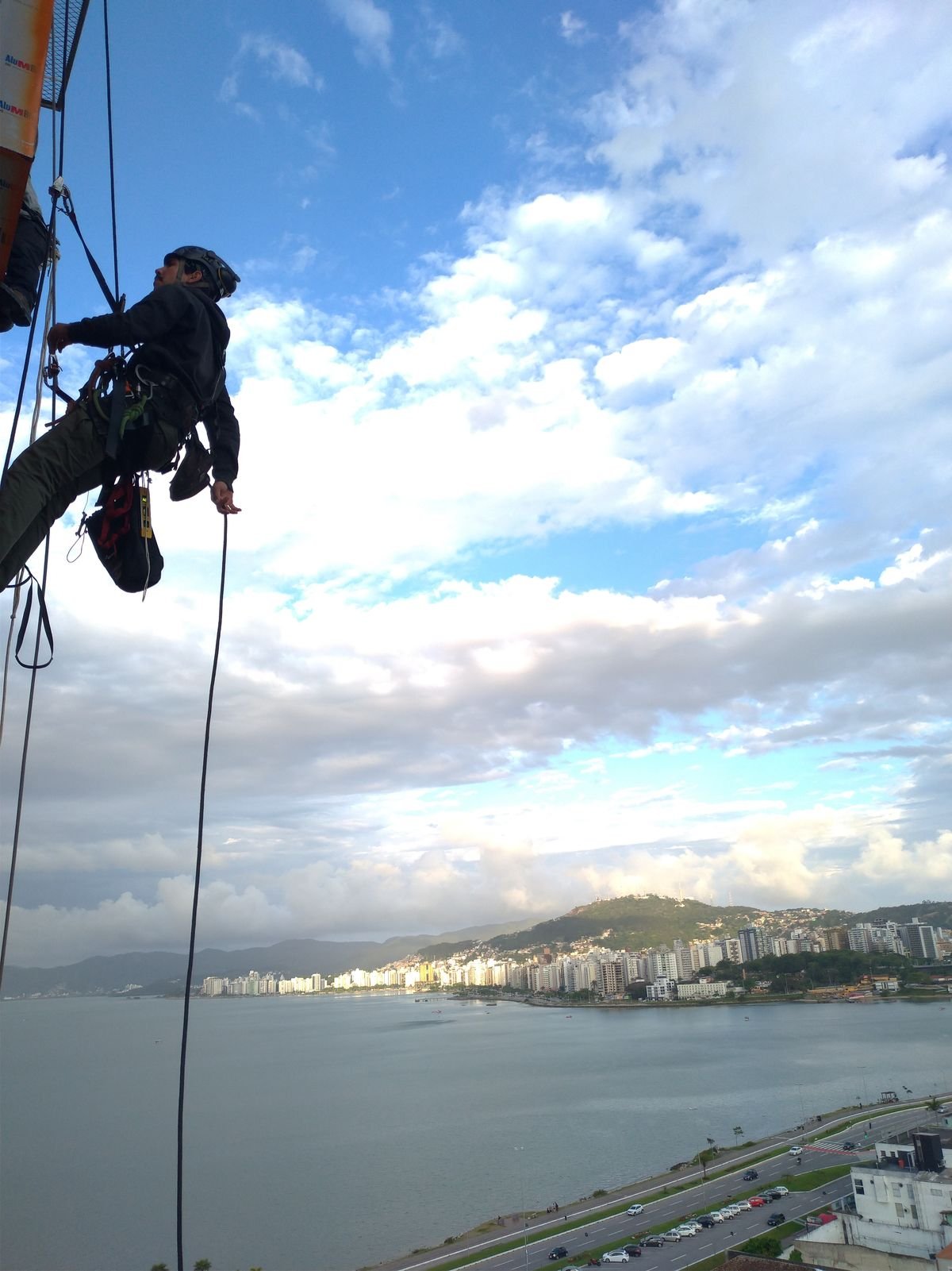 Profissional suspenso por cordas em fachada de edifício alto, com a baía de Florianópolis ao fundo